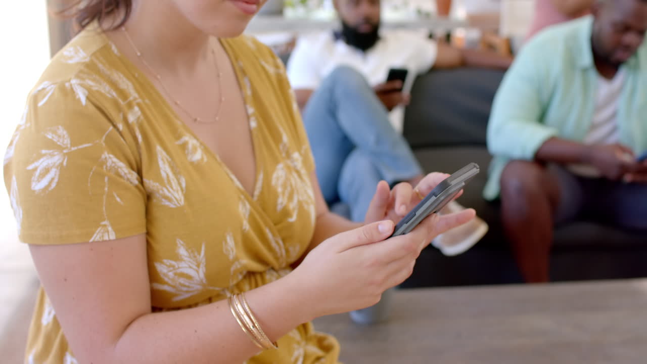 Using smartphone, woman texting while diverse friends relaxing on couch in living room