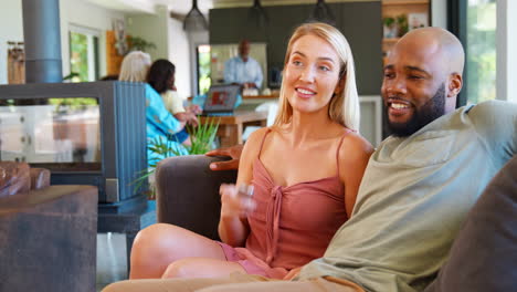 Pareja-Multirracial-En-El-Sofá-De-Casa-Juntos-Viendo-Televisión-Con-Una-Familia-Multigeneracional-En-Segundo-Plano