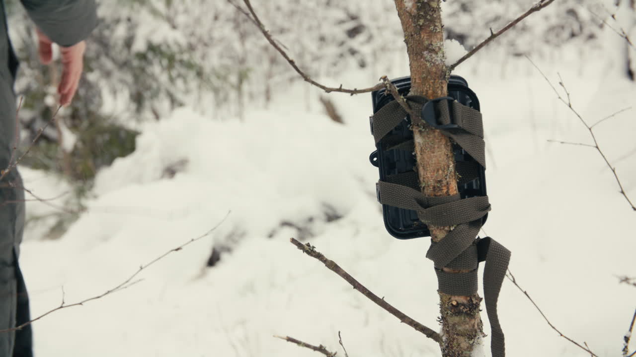 Halvnært billede af en person, der går væk gennem dyb sne efter at have opsat en vildtkamerafælde, med lav dybdeskarphed, vinterlige skovteksturer og dæmpede farver.