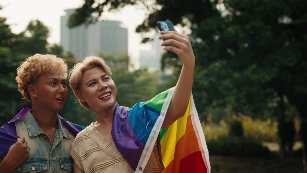 Two friends celebrate LGBTQ pride in the park, taking a selfie with the rainbow flag