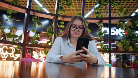 young woman seated in decorative wooden sit-out, operating phone, with shopping bags beside her, in a bustling mall restaurant, exuding a sober mood, with people moving in the background