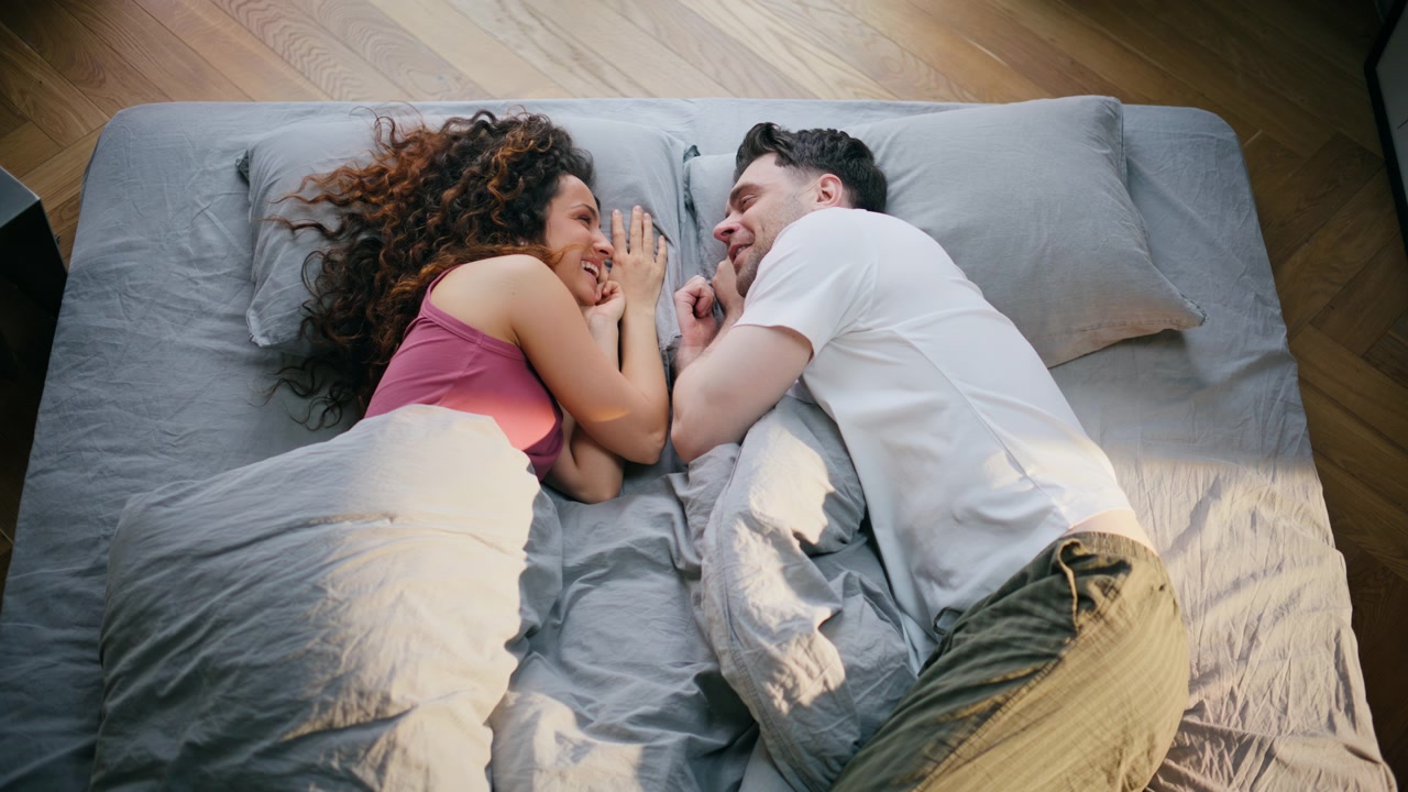Koppel van bovenaf gezien, liggend in bed, handen aanrakend. Liefdevol stel gezicht aan gezicht in de slaapkamer