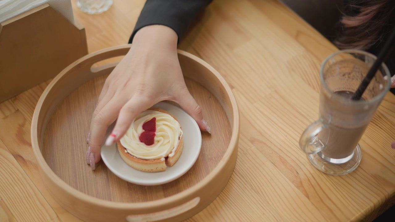 Top down view of two hands serving layered dessert beside iced coffee on wooden tabletop, showing gentle lifting, placing, and arranging in casual cafe setting during relaxed snack break