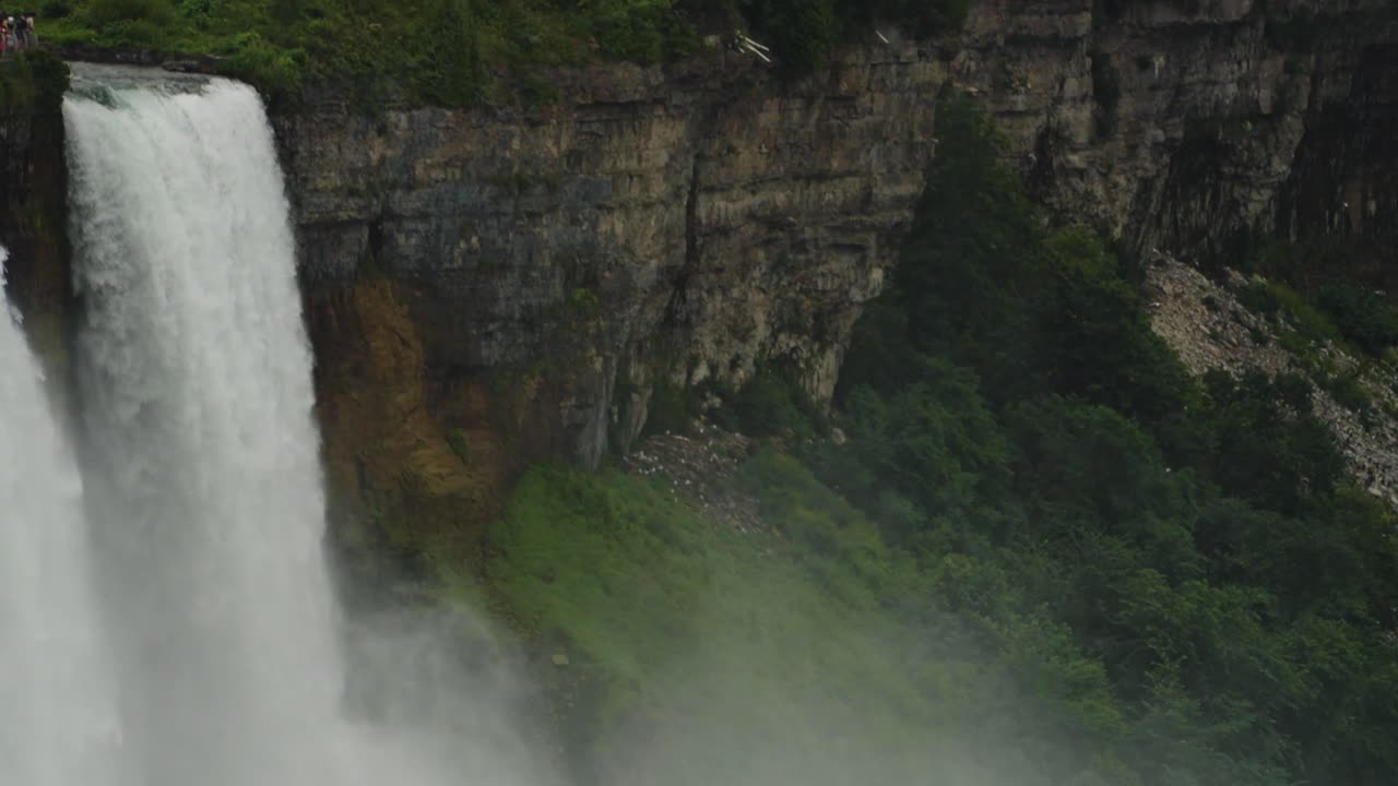 Niagara Falls plunges powerfully beside a weathered rock cliff layered in history and surrounded by lush greenery