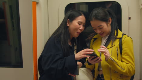 Two-Young-Female-Friends-Standing-In-Carriage-On-Underground-Train-Looking-At-Mobile-Phones