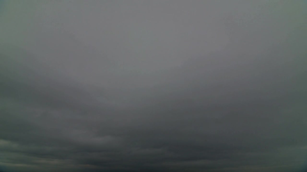 Menampilkan lapisan awan stratiform abu-abu yang perlahan bergeser di atas langit terbuka, menandakan gerimis ringan.