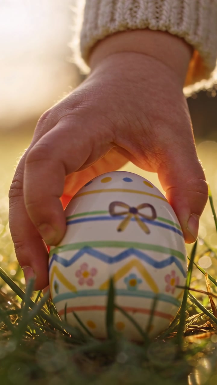 Child hand reaching, grasping and lifting painted egg from grass, revealing stripes for craft