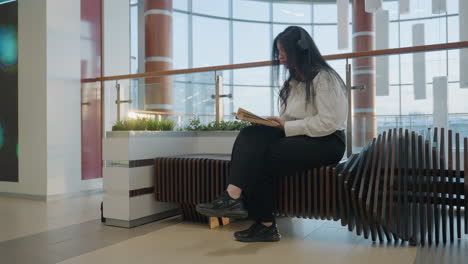 focused woman with crossed leg reads book while seated on sculptural wooden bench in modern public indoor space, wearing headphones, surrounded by architectural glass and warm natural light elements