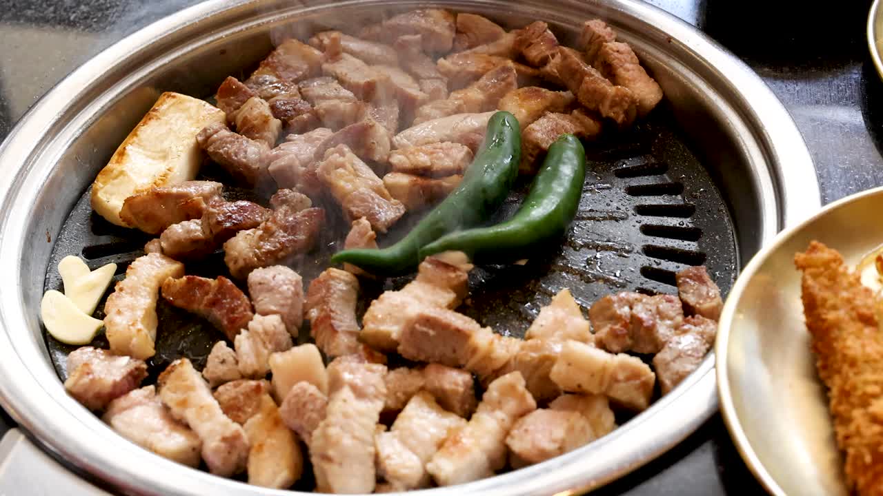Du lard de porc et des légumes grésillent sur un gril de table, des pinces ajustant les ingrédients, éclairage naturel.