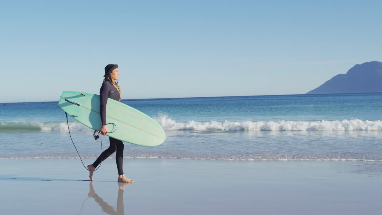 video de un hombre caucásico con rastas en traje de neopreno llevando una tabla de surf caminando por una playa soleada