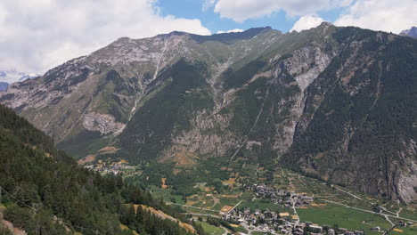 Vista-Aérea-De-Los-Alpes-Italianos-Y-Una-Pequeña-Ciudad-En-Un-Valle-Pintoresco-En-Un-Día-Soleado-De-Verano