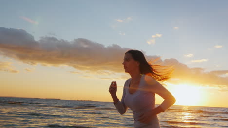 Joven-Deportista-Corre-Con-Una-Buena-Figura-En-La-Playa-Al-Atardecer-Hermoso-Cabello-Revoloteando-En-El-Wi