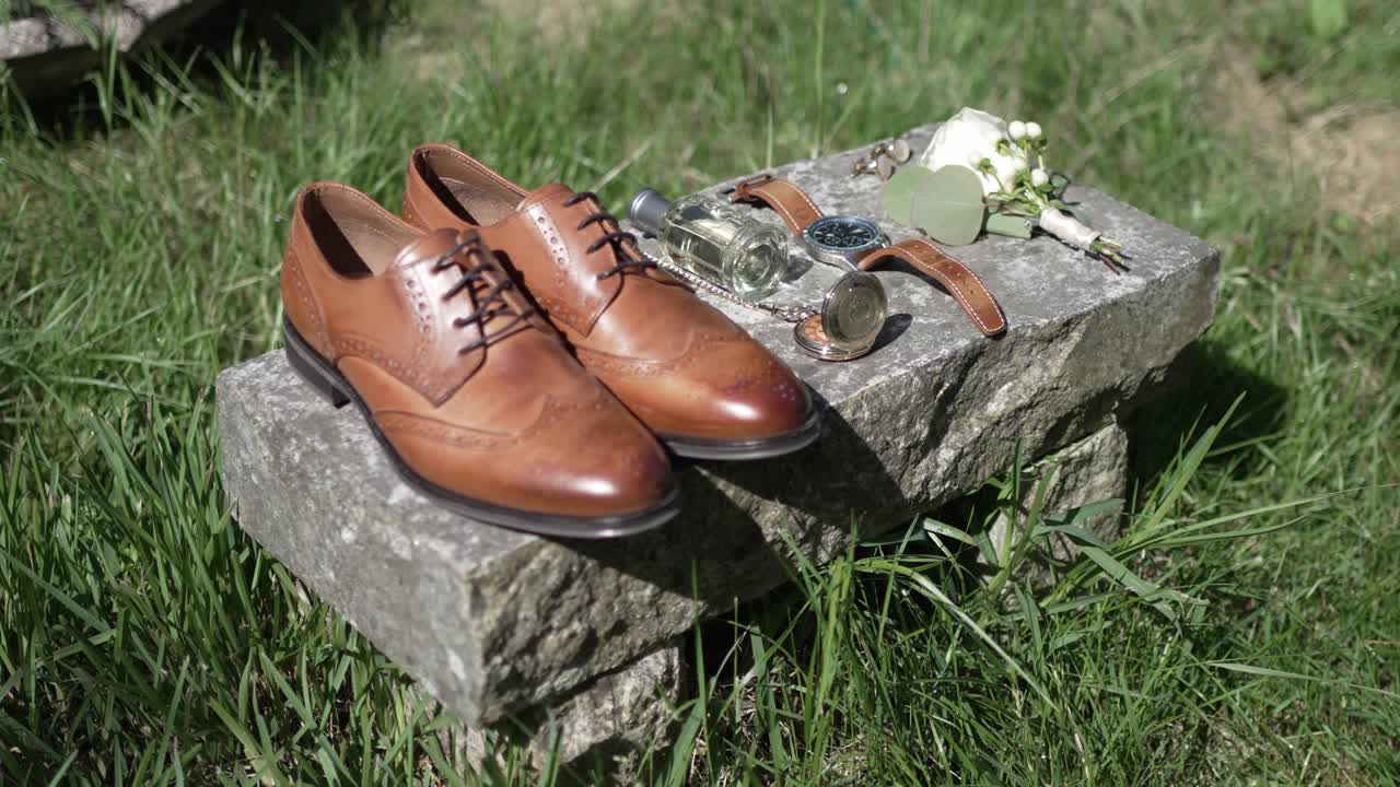 Tan leather shoes, pocket watch, sunglasses, and boutonniere arranged on stone with grass background