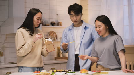 tres amigos japoneses comiendo comida japonesa alrededor del mostrador de la cocina, luego miran la cámara y sonríen