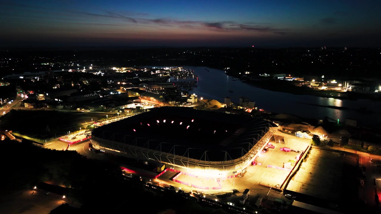 Drone aerial of the illuminated St Marys Stadium exterior and surrounding harbour lights beside the dark River Itchen in Southampton