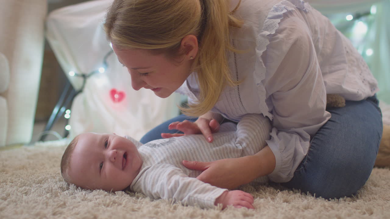 Loving mother playing game and kissing baby son lying on rug in child's bedroom at home - shot in slow motion