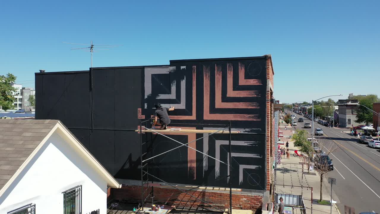 Murale dipinto a mano da un uomo su impalcatura, con vista aerea in allontanamento.