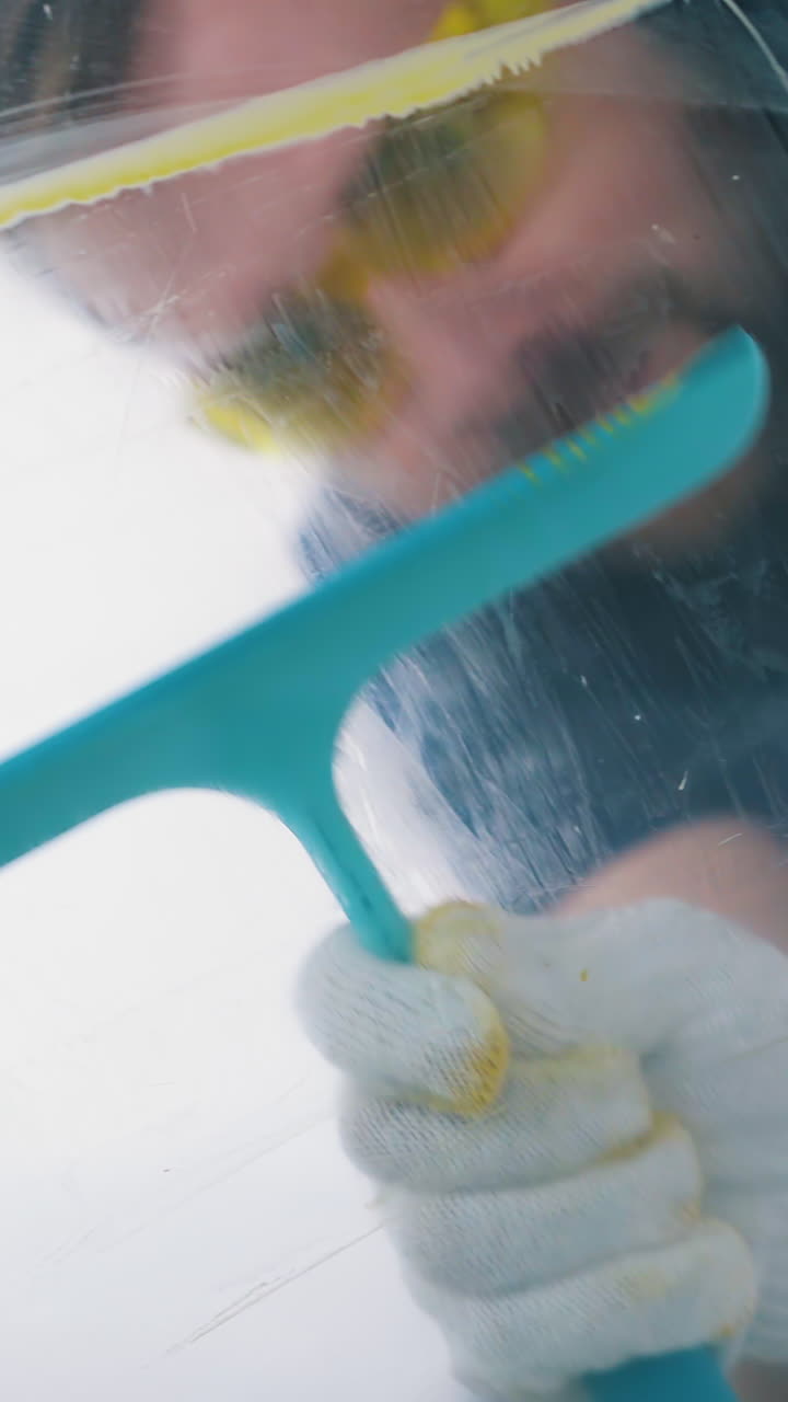 Dolly cam shot: professional builder with goggles removes paint from transparent glass with spatula at construction in light room