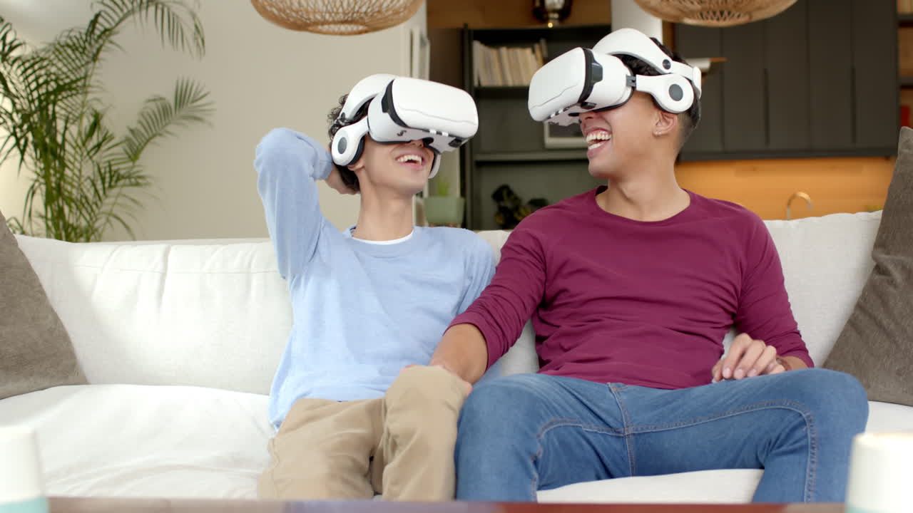 Wearing VR headsets, multiracial gay couple sitting on couch and laughing , at home