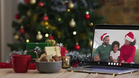 familia feliz usando sombreros snata en videollamada en computadora portátil, con decoraciones de navidad y árbol