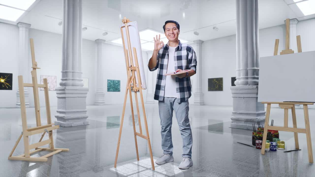 Full Body Of Asian Male Artist Smiling And Showing Okay Gesture To Camera While Painting On Canvas By Oil Paints And Brush In Gallery