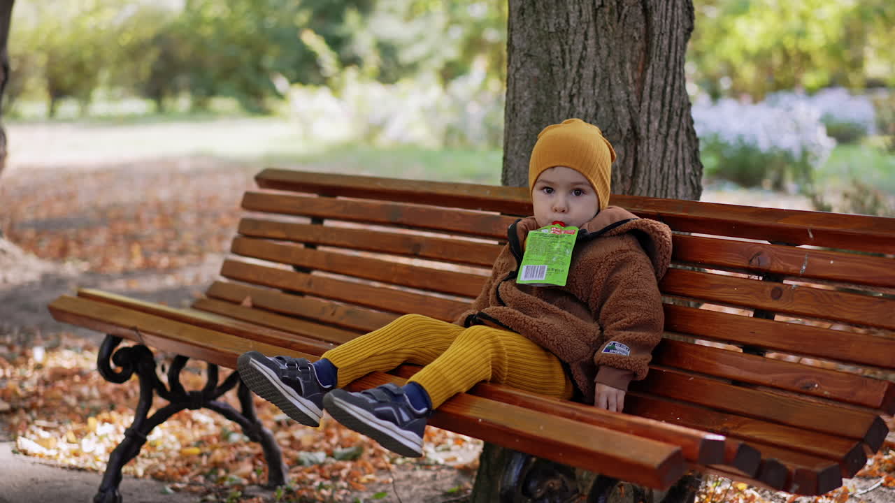Kid sits on the wooden bench in the park. Little boy holds the doypack with fruit puree in his mouth.