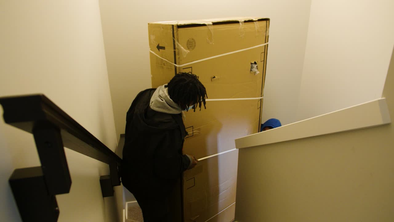 Static shot of two people carrying a heavy cardboard box up indoor stairs