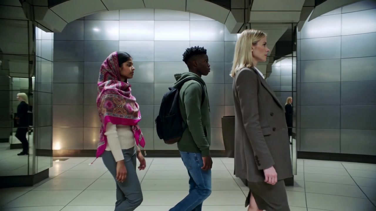 Ein gehendes Trio folgt einer Blondine im Blazer, die einen Bahnhof betritt, pendelnd mit Aktentasche und rosa Schal.