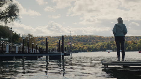 a woman stands on a pier overlooking lake ontario, the american flag is visible in the distance, there are no other people and yachts around. autumn and end of tourist season.