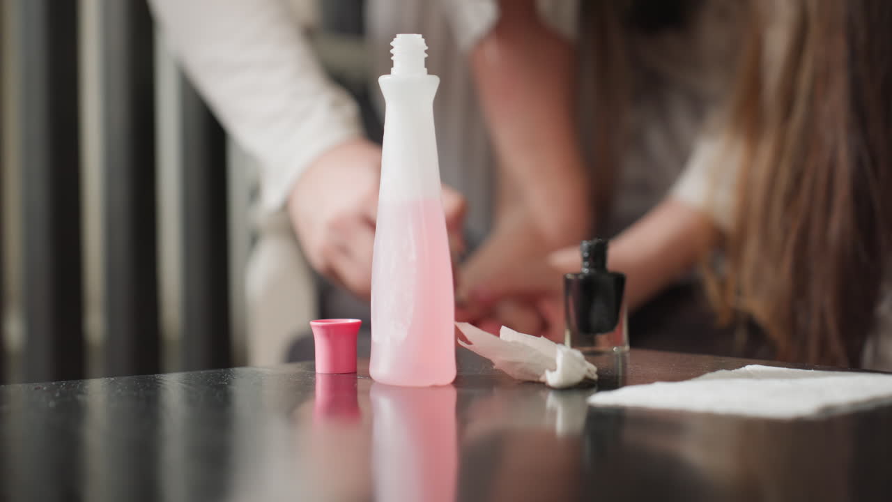 Close view of teenage girl with foot on table rubbing black polish on toes, remover bottle with open cap and tissue beside, stained wipe on surface, blurred divider, soft daylight, calm focus