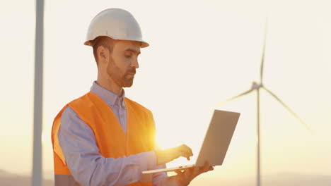 Caucasian-male-engineer-in-helmet-and-uniform-using-laptop-at-wind-station-of-renewable-energy