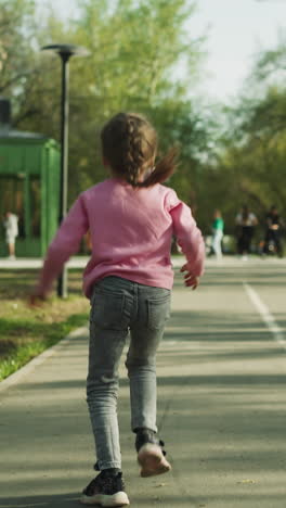 happy little girl with plait runs waving hands along road in spring city park backside view. joyful child enjoys nice weather in public garden slow motion