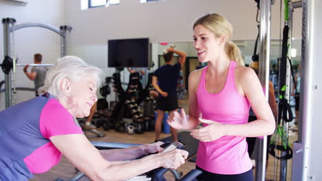 senior woman exercising on cycling machine being encouraged by personal trainer in gym