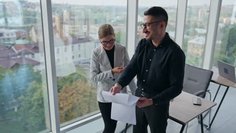 positive relaxed colleagues talking and laughing in office. man is turning the pages of documents he's holding in hands. cityscape in window at backdrop.