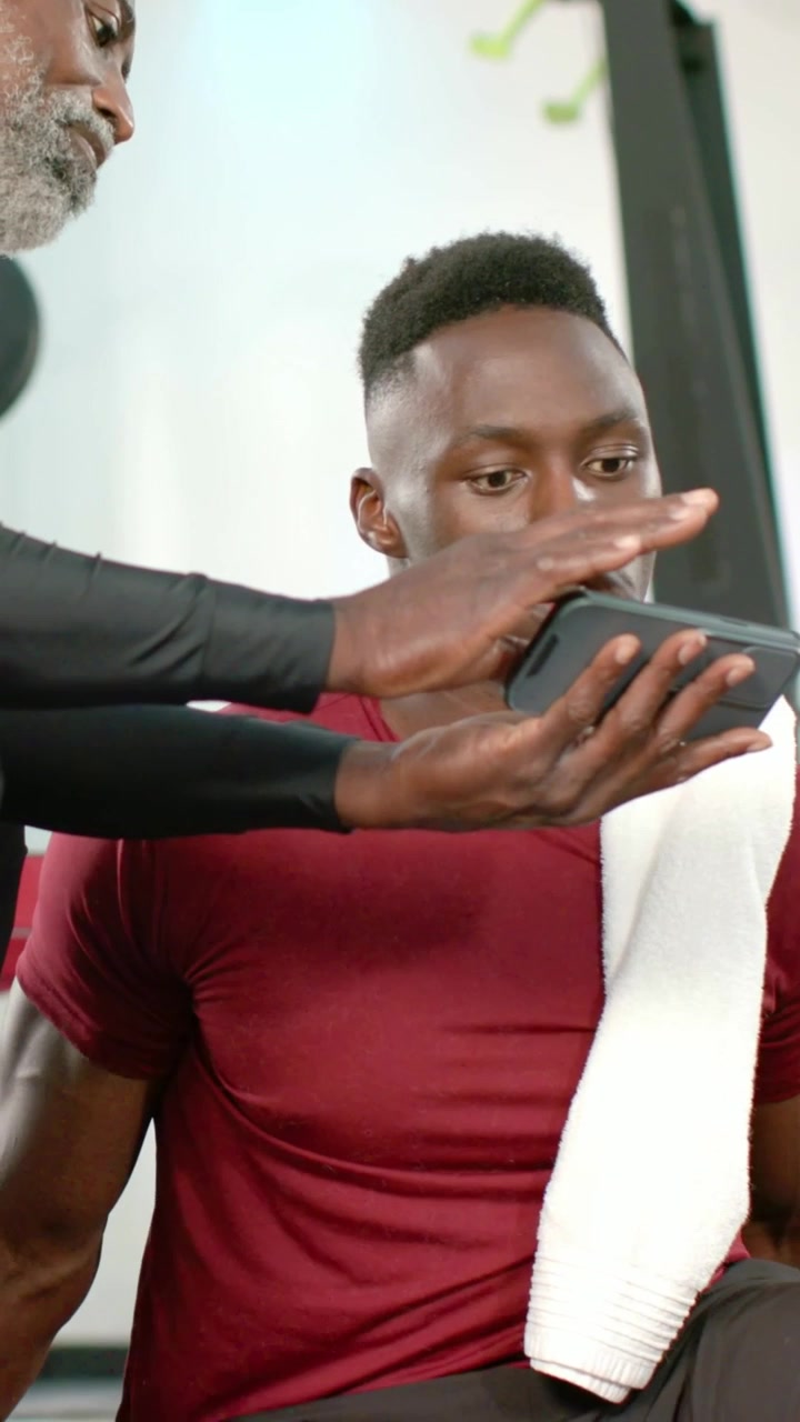 Vertical video: Showing phone, trainer tapping and teaching trainee in red shirt with towel at gym