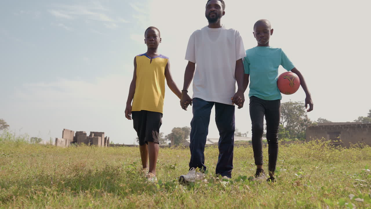 Black father standing with two sons holding basketball, front view on open grass field, confident poses, village houses in background, mentorship and playful anticipation before game