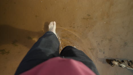 First person point of view of man's bare feet walking through a sandy creek to the other side