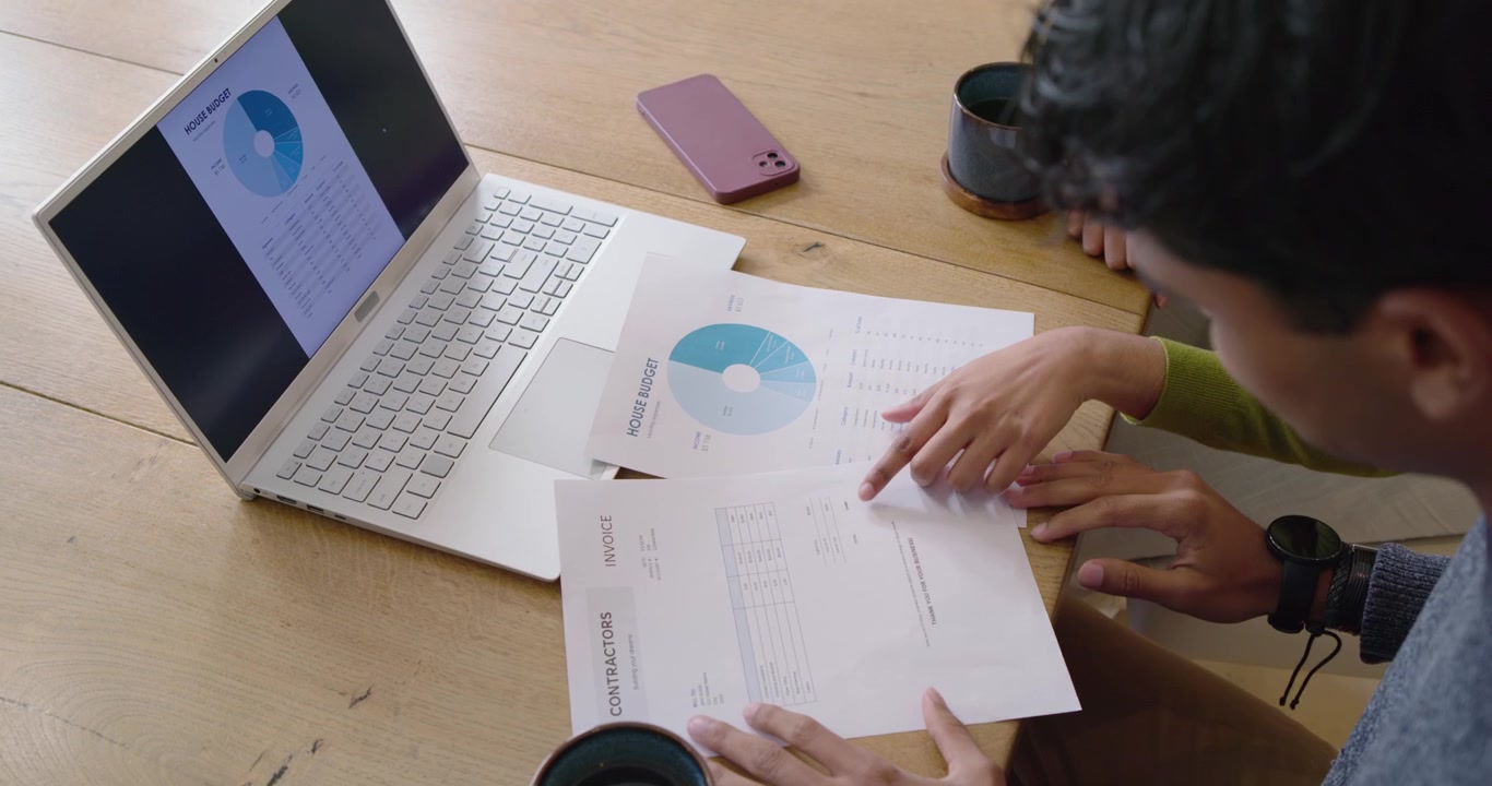 Diverse coworkers man reaching aligning laptop chart with bills on table checking totals