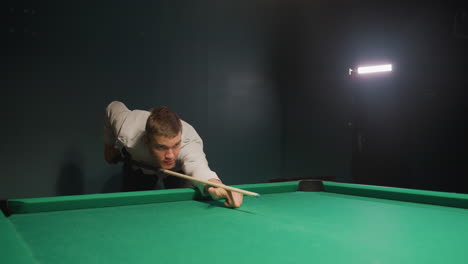 joven con camisa clara y pantalones oscuros se inclina sobre la mesa de billar verde, agarrando el palo de la pilla con intensa concentración. los ojos cerrados en la pelota de pilla mientras rueda en el bolsillo