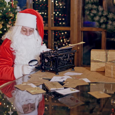 old bearded santa claus in red costume working at a table. glass table full of presents and envelopes. santa with glasses is typing letters on christmas tree background.