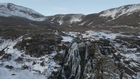 Montañas-Cubiertas-De-Nieve-Y-Cascada-Del-Velo-De-La-Novia-En-Skye-Con-Agua-En-Cascada-Y-Terreno-Accidentado,-Vista-Aérea