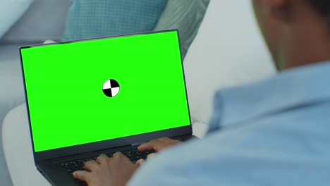 hombre étnico de raza mixta acostado en el sofá en casa, usa una computadora portátil con pantalla de maqueta verde. sobre el hombro del hombre en la sala de estar.