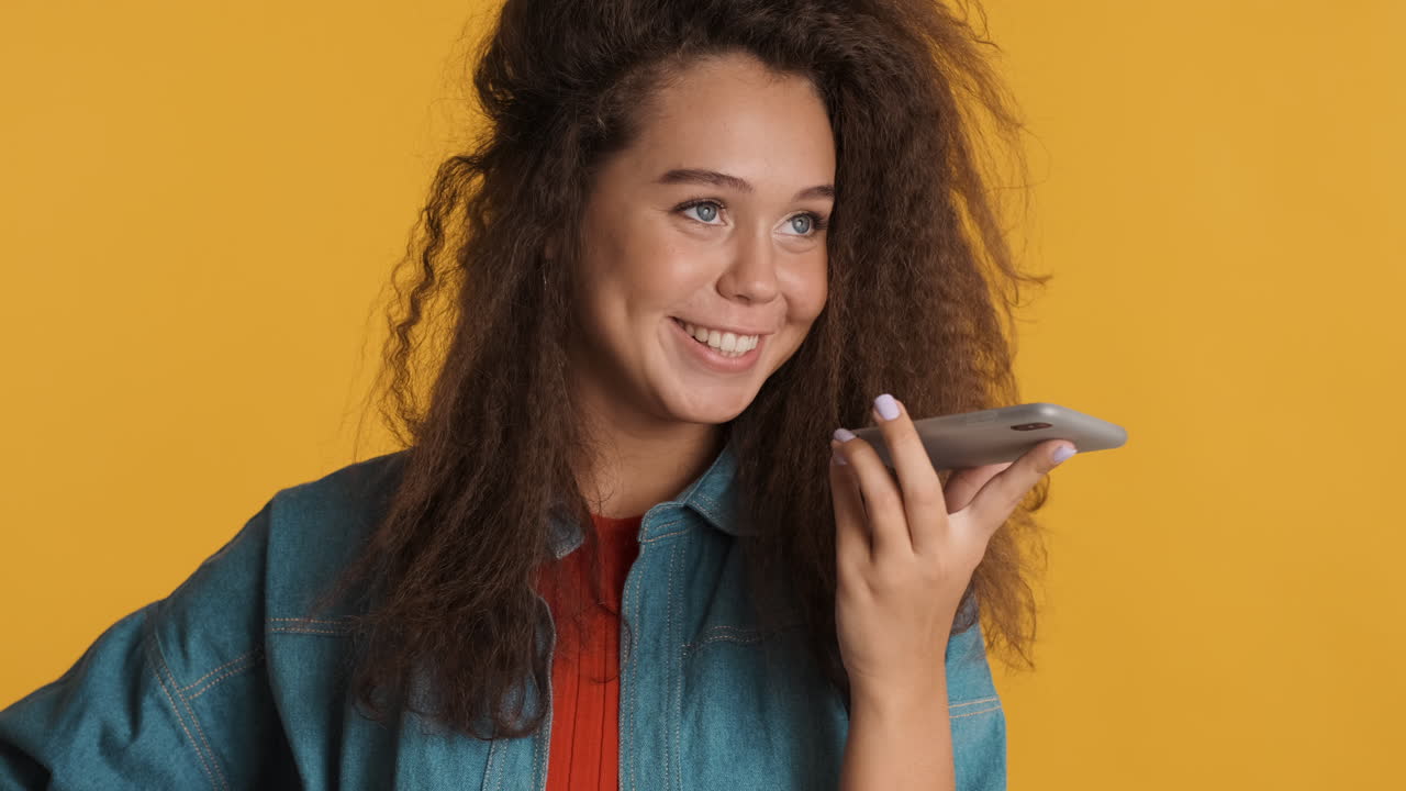 una mujer caucásica de cabello rizado grabando un mensaje de voz en su teléfono inteligente.