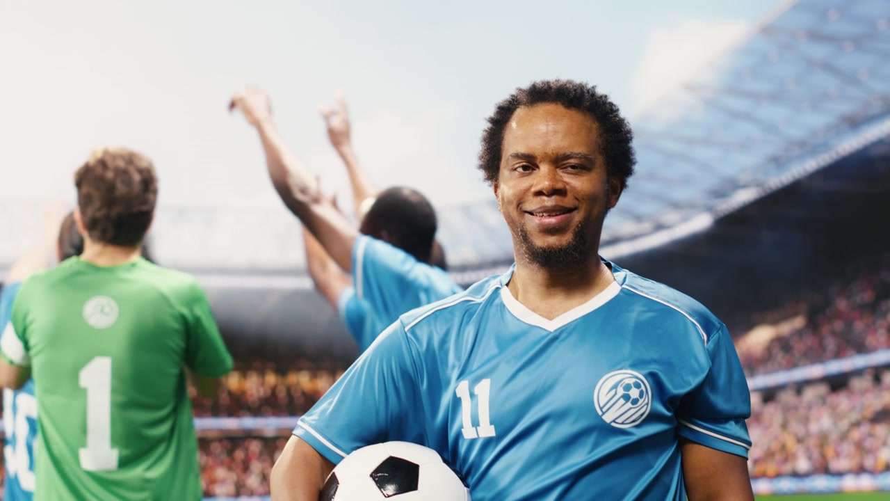 Ritratto di calciatore di colore con la maglia blu che tiene il pallone sul campo.
