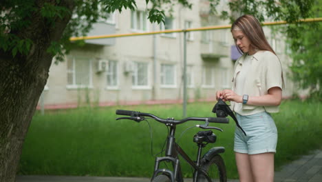 young woman stands close to her black bicycle, puts on her gloves in a peaceful urban setting, background features a residential building and yellow pole, surrounded by greenery