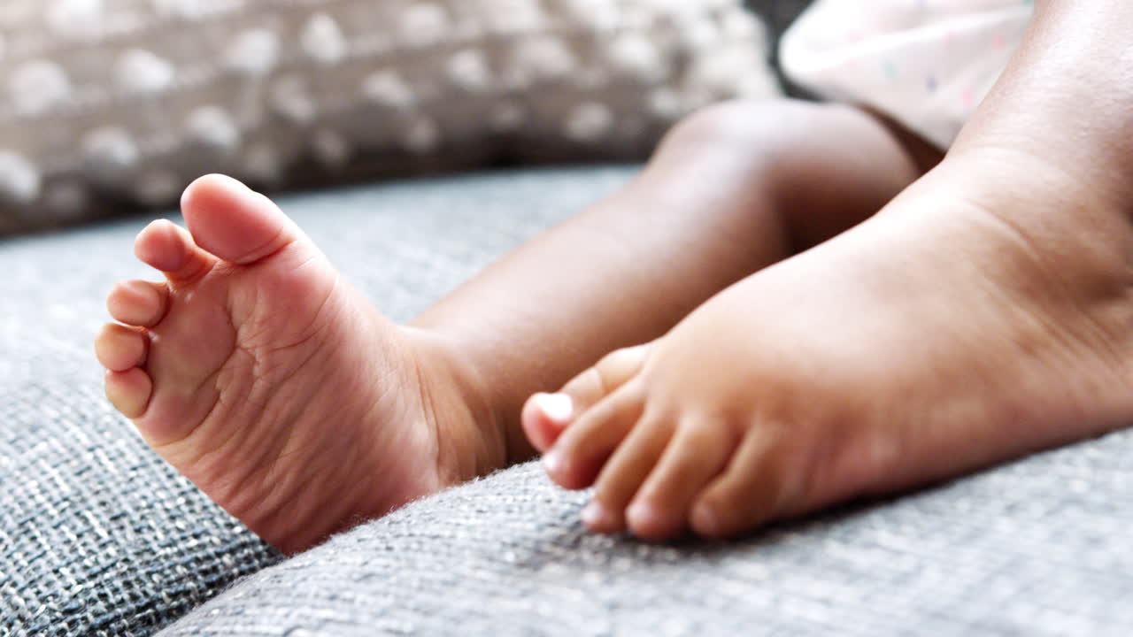 Close Up Of Baby Girl Sitting On Sofa At Home Playing With Toes