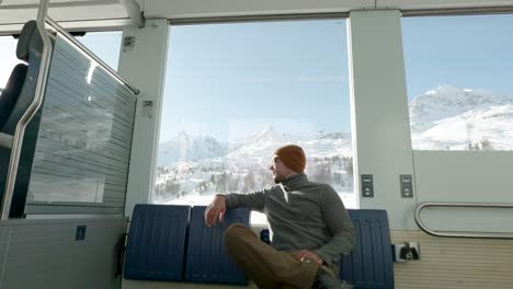 Young-caucasian-man-sitting-next-to-a-train-window-and-looking-outside-to-snow-covered-mountains-during-a-sunny-winter-day