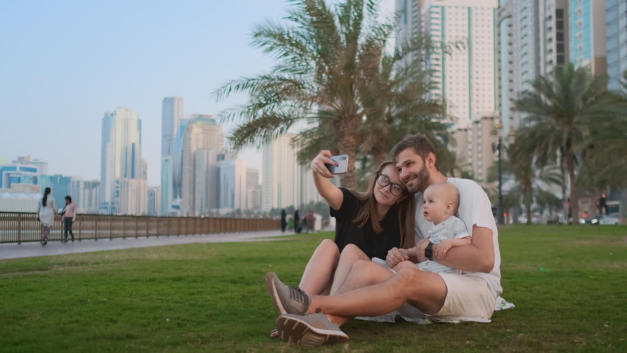 Happy family with two children sitting together on grass in park and taking a selfie. With smartphone.