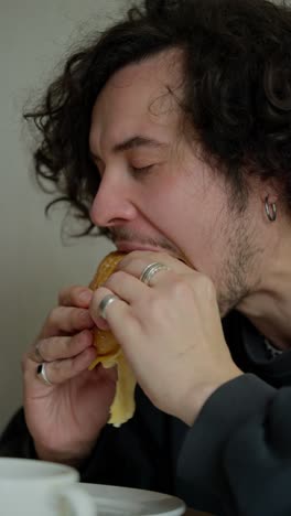 vertical video close up of a sleepy brunette guy with a mustache and curly hair eating his sandwich in the morning and enjoying it