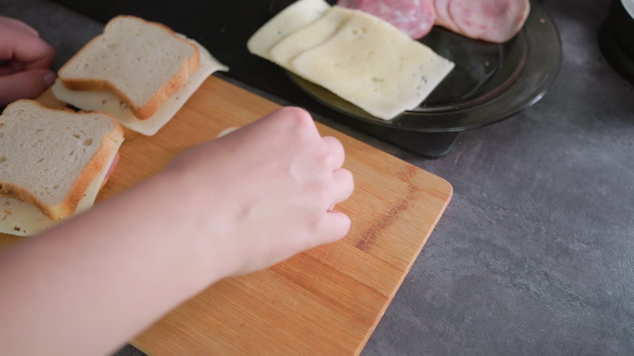Closeup of hand picking cold cuts from black plate while preparing sandwich with sliced white bread and cheese on wooden cutting board during homemade meal preparation in clean modern kitchen setting